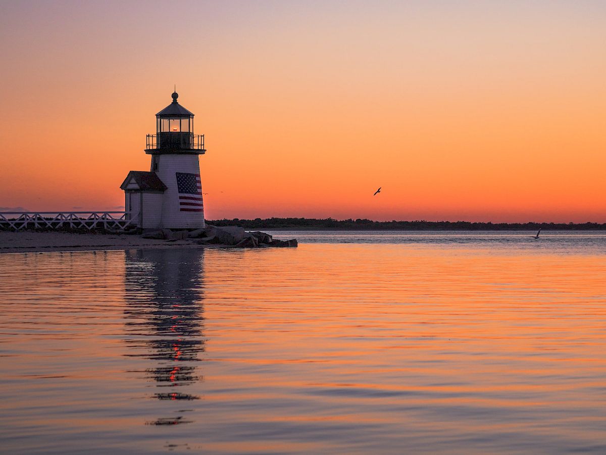 A lighthouse standing by a calm sea at sunset, with an orange and purple sky reflecting on the water, and a single bird flying in the distance.