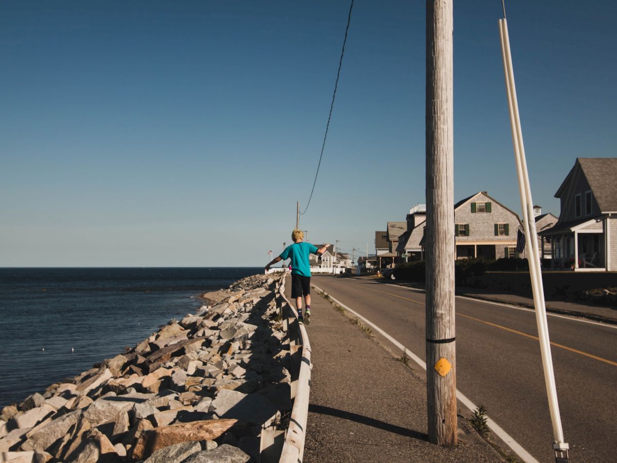 A person in a blue shirt and hat walks along a rocky seawall next to a road with houses nearby and a telephone pole in the foreground.