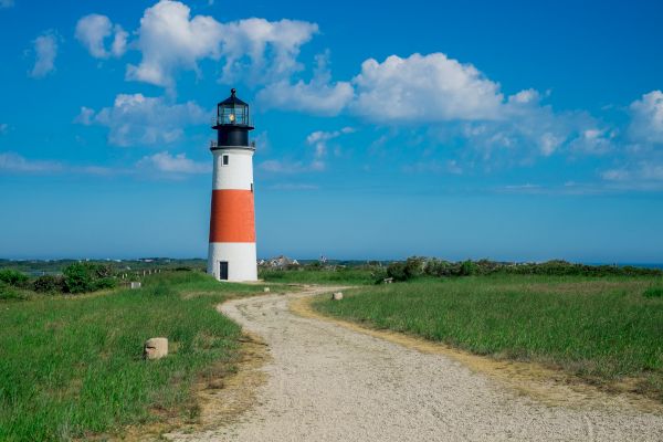 A lighthouse with red and white stripes stands on a grassy landscape under a blue sky with clouds, with a dirt path leading to it, ending the sentence.