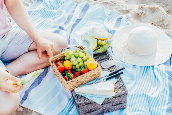 A beach picnic scene with a person, a basket of fruits, a hat, and a striped mat. The basket contains grapes, strawberries, and other fruits.